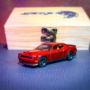 May include: A metallic red toy car model of a Dodge Challenger with black wheels and a black rear spoiler. The car features a detailed front grille, hood vents, and a visible interior. Behind the car sits a light-colored wooden box with a metal latch.