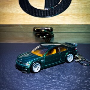 May include: A dark green die-cast model car with white wheels and a keychain. The car has a yellow interior and is set against a wooden box with a BMW logo. The car is a miniature replica of a sports car.