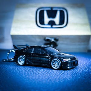 Puede incluir: Un coche de juguete negro con alerón y ruedas plateadas, sujeto a un llavero. El coche está frente a una caja de madera con el logotipo de Honda. El fondo es una superficie azul texturizada.