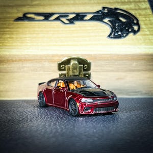 Puede incluir: Un coche de juguete rojo Dodge Charger SRT Hellcat con capó negro y ruedas negras. El coche está sobre una superficie negra.