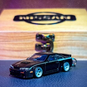 May include: A black die-cast model car keychain featuring a coupe design with light blue wheels, positioned in front of a light-colored wooden box with a black Nissan logo printed on the lid and a metal latch closure on the front.