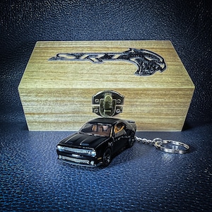 Puede incluir: Un llavero negro Dodge Challenger SRT con una caja de madera. El llavero presenta una réplica detallada del coche con un acabado negro y un anillo de llave plateado.