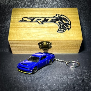 Puede incluir: Un llavero Dodge Challenger SRT azul con una caja de madera. La caja tiene el logotipo SRT y una cabeza de lobo grabada en la parte superior.
