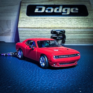 Puede incluir: Un coche modelo Dodge Challenger rojo con llantas plateadas e interior negro. El coche está sobre una superficie azul oscuro, con una caja de madera y el logotipo de Dodge en el fondo.