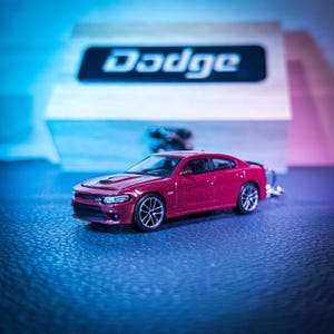 Puede incluir: Un coche modelo Dodge Charger rojo con ruedas negras y un diseño detallado. El coche está sobre un fondo borroso con el logotipo de Dodge en una caja de madera. La imagen está iluminada con tonos azules y rosas.