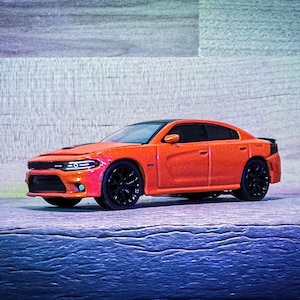 Puede incluir: Un coche de juguete naranja con ruedas negras y techo negro. El coche es un sedán, colocado sobre una superficie azul y gris. La parte delantera del coche presenta faros y una parrilla detallados.