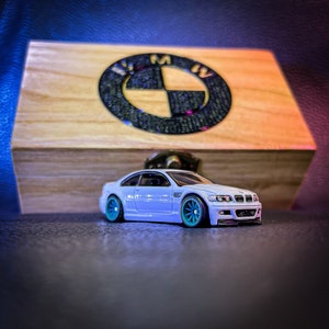 May include: A white toy car with teal wheels is in front of a wooden box with a BMW logo. The car is a coupe with a detailed design. The box has a light wood finish and a dark logo.