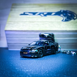 Puede incluir: Un coche de juguete negro con la palabra "HELLCAT" en la parte delantera, sujeto a un llavero. El coche está sobre una superficie azul, con una caja de madera y un diseño de llave en el fondo.
