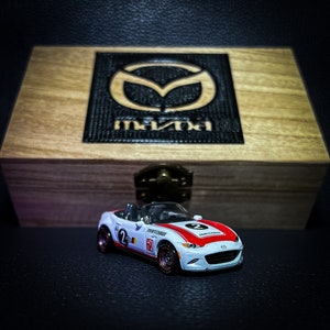 Puede incluir: Un coche de juguete blanco y rojo con la palabra "Mazda" en el lateral. El coche está encima de una caja de madera con el logotipo de Mazda en la parte superior.