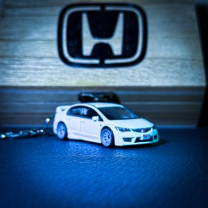 May include: A white model car with blue wheels and a keychain. The car is in front of a wooden surface with a Honda logo. The car is a sedan style.