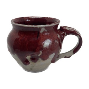 May include: A handmade ceramic mug with a unique, drippy glaze in shades of red and white. The mug has a rounded body and a comfortable handle.