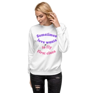 Puede incluir: Sudadera blanca con el texto "Sometimes love wants to fly first class" en letras moradas y rosas. Un pequeño icono de avión está por encima de la palabra "fly".