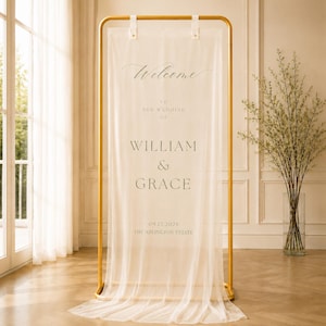 May include: A tall, rectangular gold metal frame holding a sheer, off-white fabric wedding sign. The text reads: Welcome to the wedding of William & Grace, 09.12.2026, The Arlington Estate. The fabric drapes elegantly to the floor.