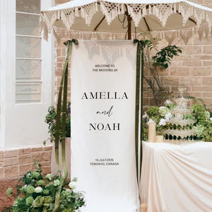 Peut inclure: Un grand panneau de bienvenue de mariage rectangulaire blanc avec le texte noir : WELCOME TO THE WEDDING OF AMELLA and NOAH 13.JULY.2025 TORONTO, CANADA. Le panneau est orné de longs rubans vert foncé attachés aux coins supérieurs.