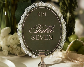 May include: An oval table number card in dark green with rose gold text reading C|M, Table SEVEN, and OCTOBER 9TH 2026. The card is bordered by white lace trim and is held upright by a small silver metal stand on a table setting.