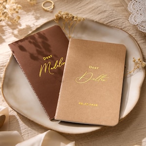 Puede incluir: Dos cuadernos rectangulares con esquinas redondeadas, uno marrón oscuro y otro marrón claro, con texto en lámina dorada. El cuaderno marrón oscuro dice Dear Madeline y el marrón claro dice Dear Dalton con la fecha 09.27.2026 debajo.
