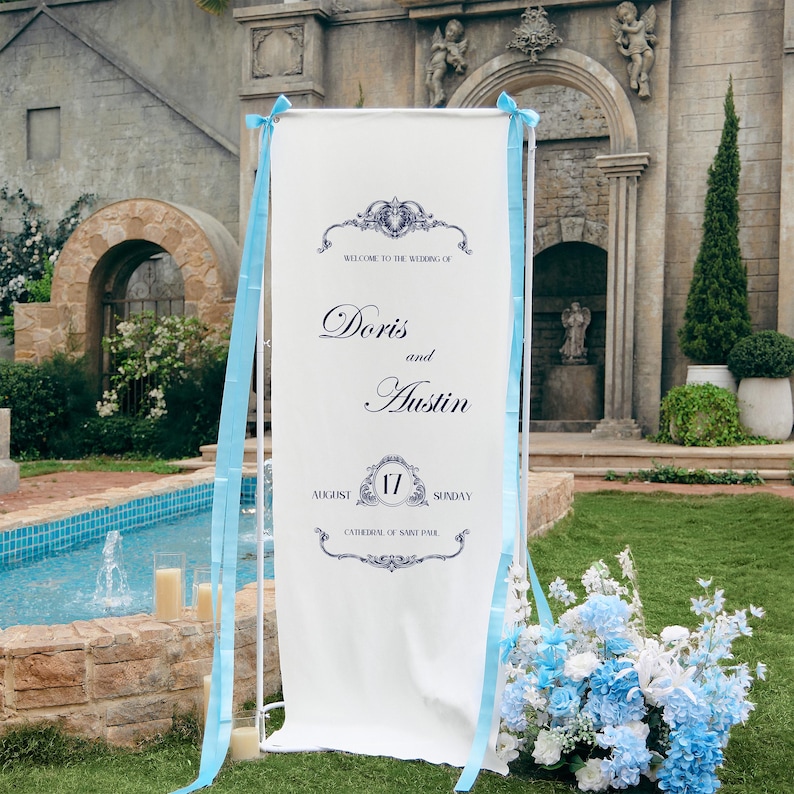 May include: A tall white rectangular wedding welcome sign on a metal stand, featuring dark blue text reading Welcome to the wedding of Doris and Austin, August 17 Sunday, Cathedral of Saint Paul, decorated with light blue ribbons on the sides.