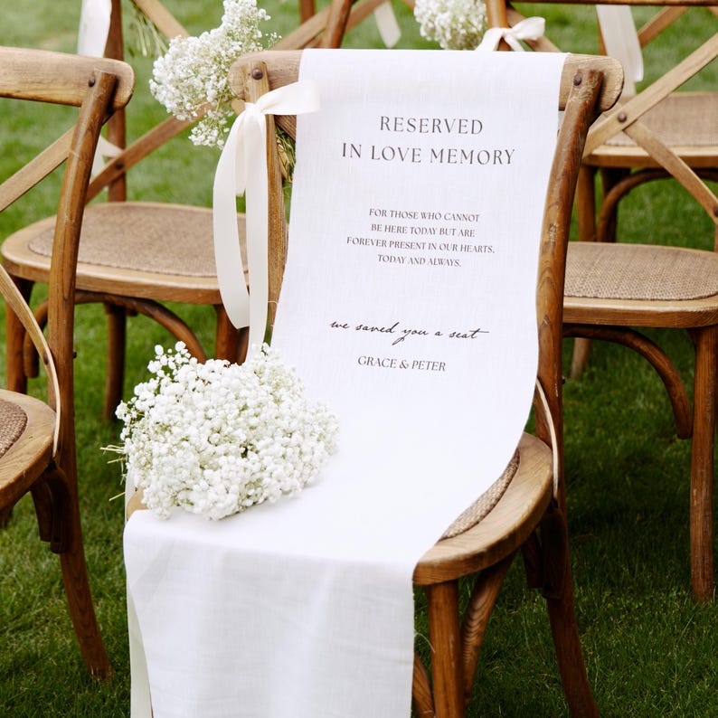 May include: A white fabric sign draped over a wooden chair, featuring the text RESERVED IN LOVE MEMORY, FOR THOSE WHO CANNOT BE HERE TODAY BUT ARE FOREVER PRESENT IN OUR HEARTS, TODAY AND ALWAYS, we saved you a seat, GRACE & PETER, with baby's breath.