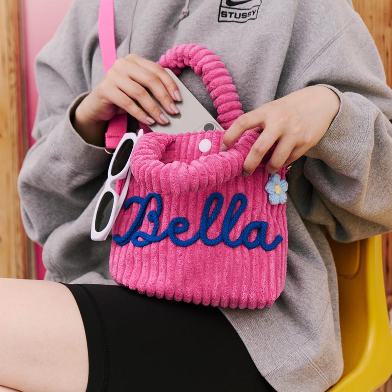 May include: A pink corduroy tote bag with the name "Bella" embroidered in blue script. The bag has a matching pink strap and handle, with a small blue flower charm. White sunglasses hang from the side.