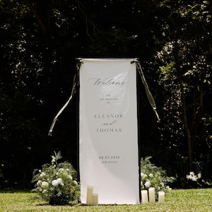 May include: A white wedding welcome sign with black text that reads "Welcome to the wedding of Eleanor and Thomas 08.07.2025 San Diego." The sign is framed with a black border and green ribbons. White candles and floral arrangements are at the base.