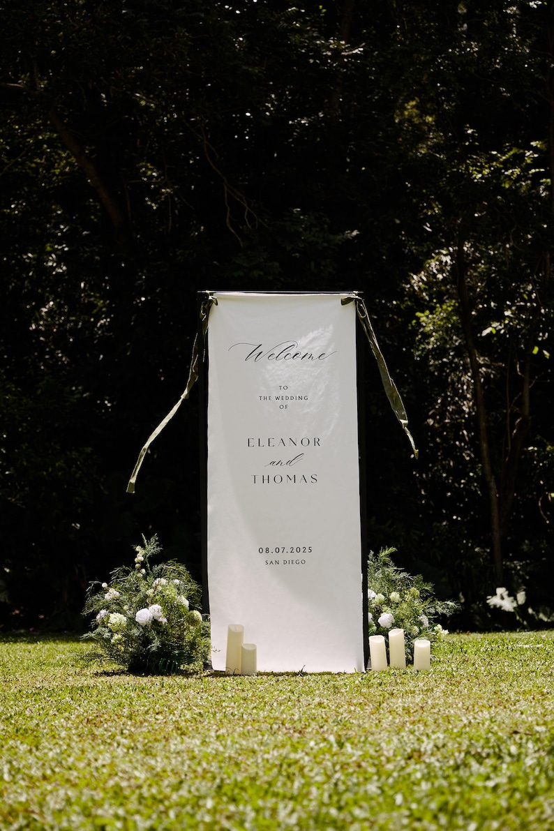 May include: A tall, rectangular white fabric wedding welcome sign suspended from a black frame with olive green ribbons. The text reads: Welcome to the wedding of Eleanor and Thomas 08.07.2025 San Diego. White candles and green floral arrangements sit at the base.