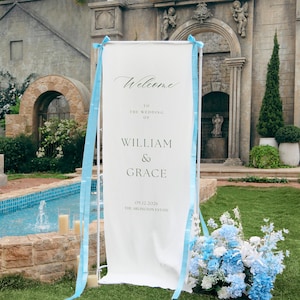 May include: A tall, rectangular white wedding welcome sign held by a white metal frame, decorated with light blue ribbons. The text reads: Welcome to the wedding of William & Grace, 09.12.2026, The Arlington Estate, in black serif font.