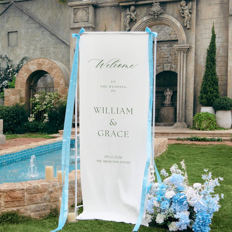 May include: A tall, rectangular white wedding welcome sign held by a white metal frame, decorated with light blue ribbons. The text reads: Welcome to the wedding of William & Grace, 09.12.2026, The Arlington Estate, in black serif font.