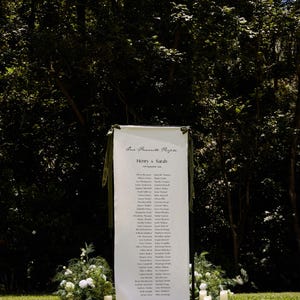 May include: A white banner with the text "Our Favorite People" and the names of guests for a wedding. The banner is decorated with green ribbons and stands on a grassy lawn. White candles and floral arrangements are placed around the banner.