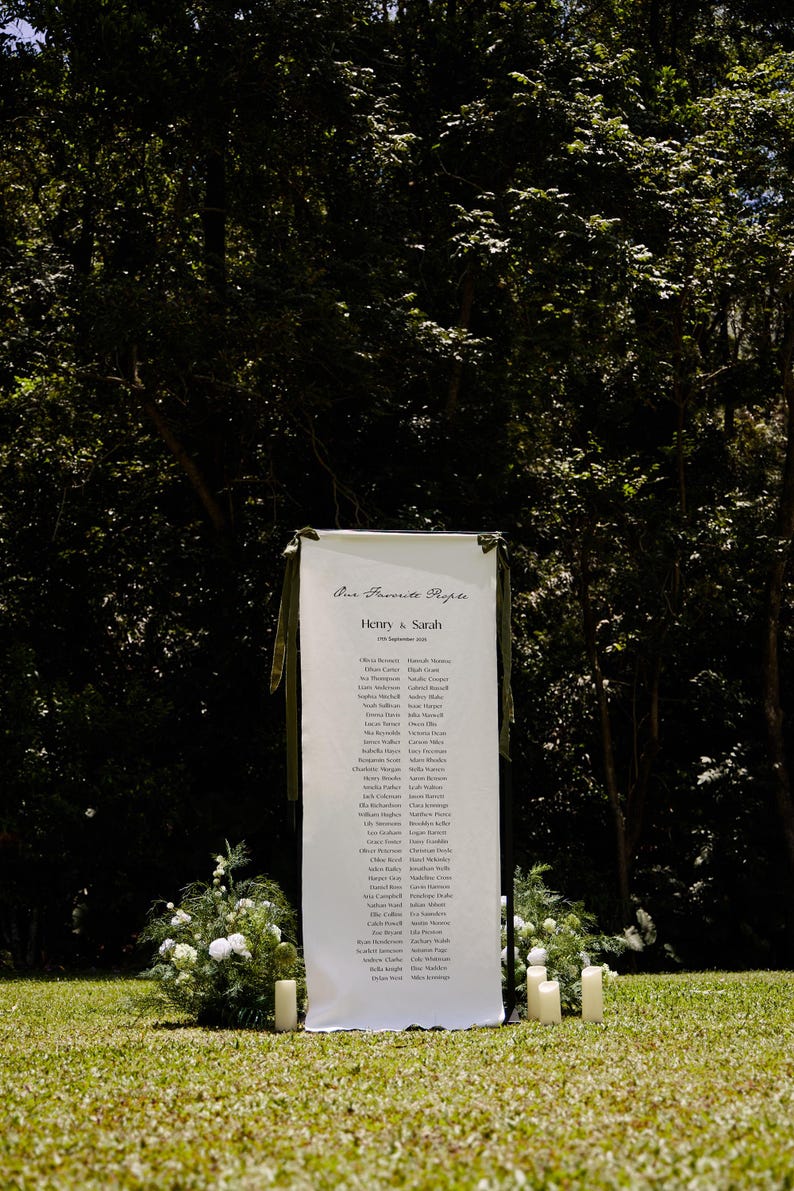 May include: A white banner with the text "Our Favorite People" and the names of guests for a wedding. The banner is decorated with green ribbons and stands on a grassy lawn. White candles and floral arrangements are placed around the banner.