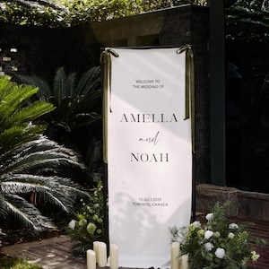 May include: A white wedding welcome sign with the text "WELCOME TO THE WEDDING OF" above the names "AMELLA and NOAH". The date "13. JULY.2025 TORONTO, CANADA" is printed below. The sign is decorated with green ribbons and surrounded by greenery and candles.