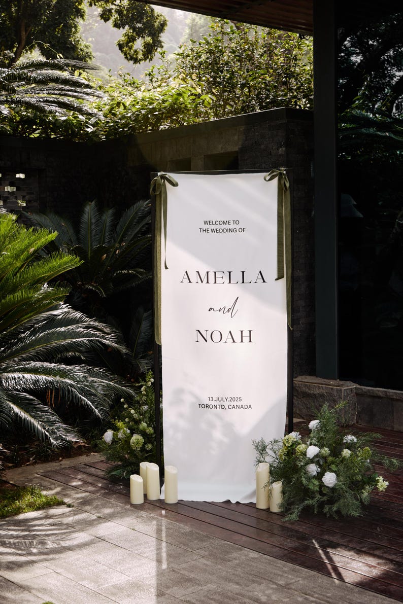 May include: A white wedding welcome sign with the text "WELCOME TO THE WEDDING OF" above the names "AMELLA and NOAH". The date "13. JULY.2025 TORONTO, CANADA" is printed below. The sign is decorated with green ribbons and surrounded by greenery and candles.