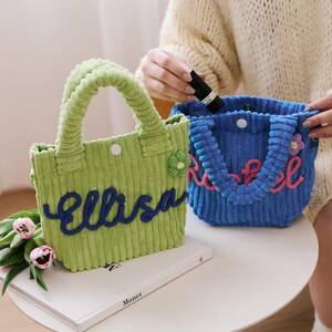 May include: Two corduroy tote bags, one green with "ellisa" in blue script and the other blue with "Rachel" in pink script. Each bag has a handle, a flower accent, and a button closure. The bags are on a white table.