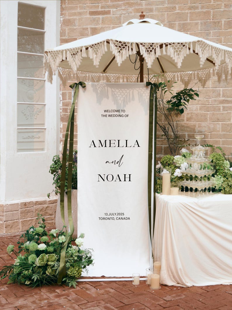 May include: A tall, rectangular white wedding welcome sign featuring black text that reads: WELCOME TO THE WEDDING OF AMELLA and NOAH 13.JULY.2025 TORONTO, CANADA. The sign is accented with long, dark green ribbons tied to the top corners.