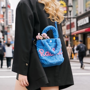 May include: A blue corduroy tote bag with the word "Race" in pink script. The bag has a top handle and is filled with pink flowers. The bag is worn over the shoulder.