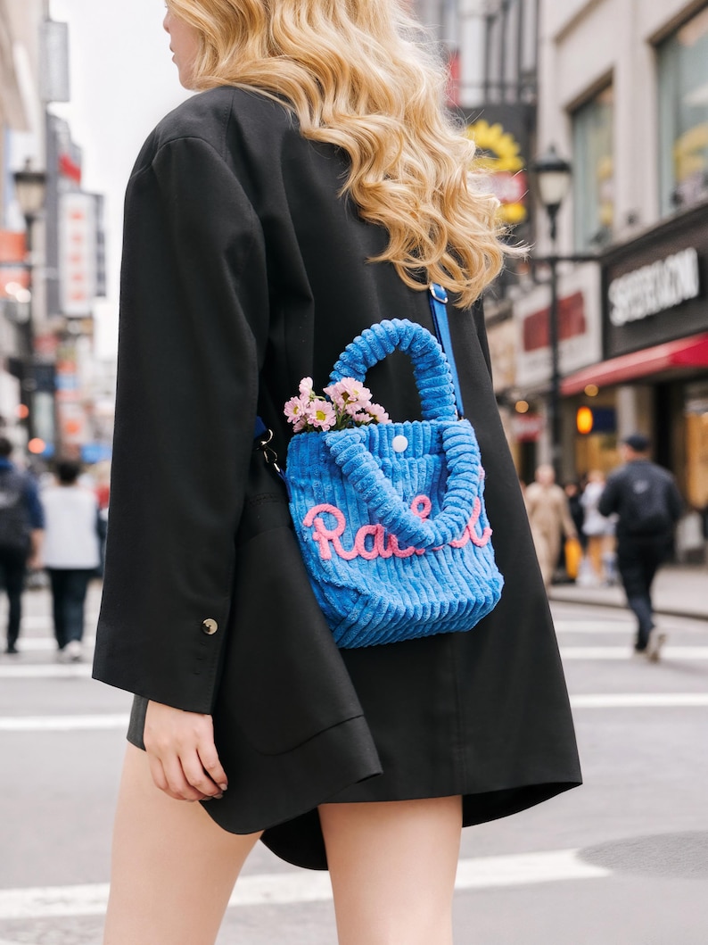 May include: A blue corduroy tote bag with the word "Race" in pink script. The bag has a top handle and is filled with pink flowers. The bag is worn over the shoulder.