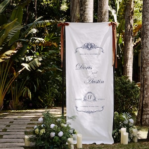 May include: A white wedding sign with brown ribbon ties, set outdoors. The sign reads "Welcome to the Wedding of Doris and Austin" with the date August 17th. Decorative floral arrangements and candles flank the sign, with a stone pathway in the foreground.