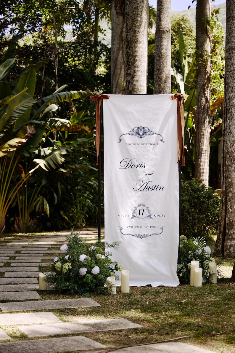 May include: A white wedding sign with brown ribbon ties, set outdoors. The sign reads "Welcome to the Wedding of Doris and Austin" with the date August 17th. Decorative floral arrangements and candles flank the sign, with a stone pathway in the foreground.