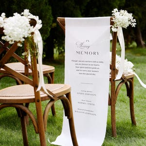 May include: Wooden chairs adorned with white flowers and a white banner reading "In Loving Memory." The banner displays names and a message. The chairs are arranged on a green lawn, indicating an outdoor memorial or wedding setting.