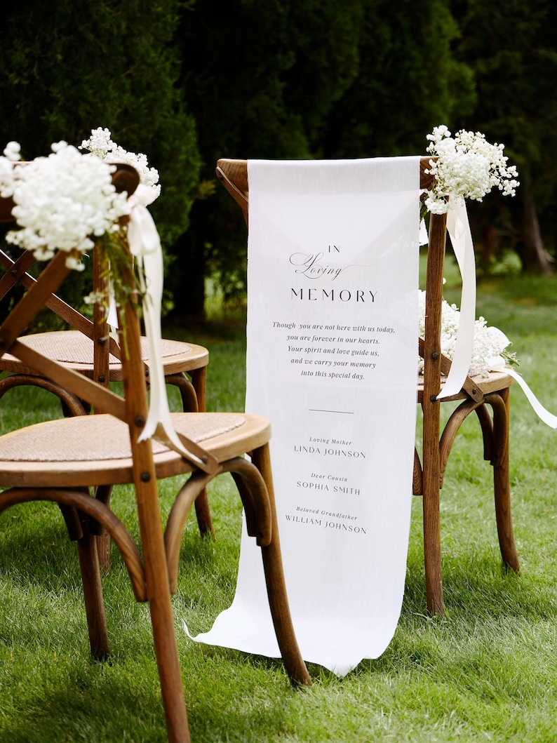 May include: Two wooden cross-back chairs with woven seats, each adorned with white baby's breath flowers and ribbons. A long white fabric banner hangs from one chair, printed with the text: IN Loving MEMORY, followed by a poem and three names.