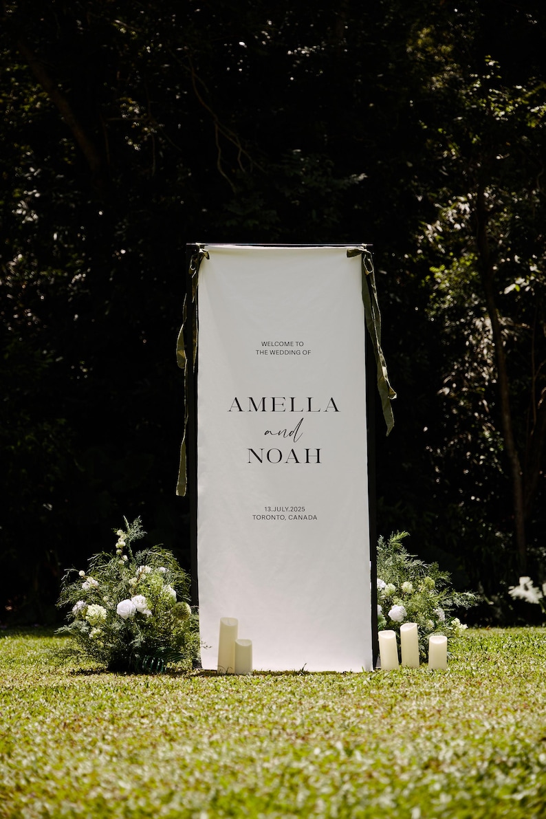 May include: A white wedding welcome sign with the names Amella and Noah, the date July 13, 2025, and the location Toronto, Canada. The sign is decorated with green ribbons and stands on a grassy lawn with floral arrangements and candles.