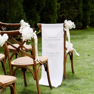 May include: A wooden cross-back chair featuring a white fabric sign draped over the backrest. The sign reads: RESERVED IN LOVING MEMORY, FOR THOSE WHO CANNOT BE HERE TODAY BUT ARE FOREVER PRESENT IN OUR HEARTS, TODAY AND ALWAYS, we saved you a seat, GRACE & PETER. White flowers are tied to the chair.