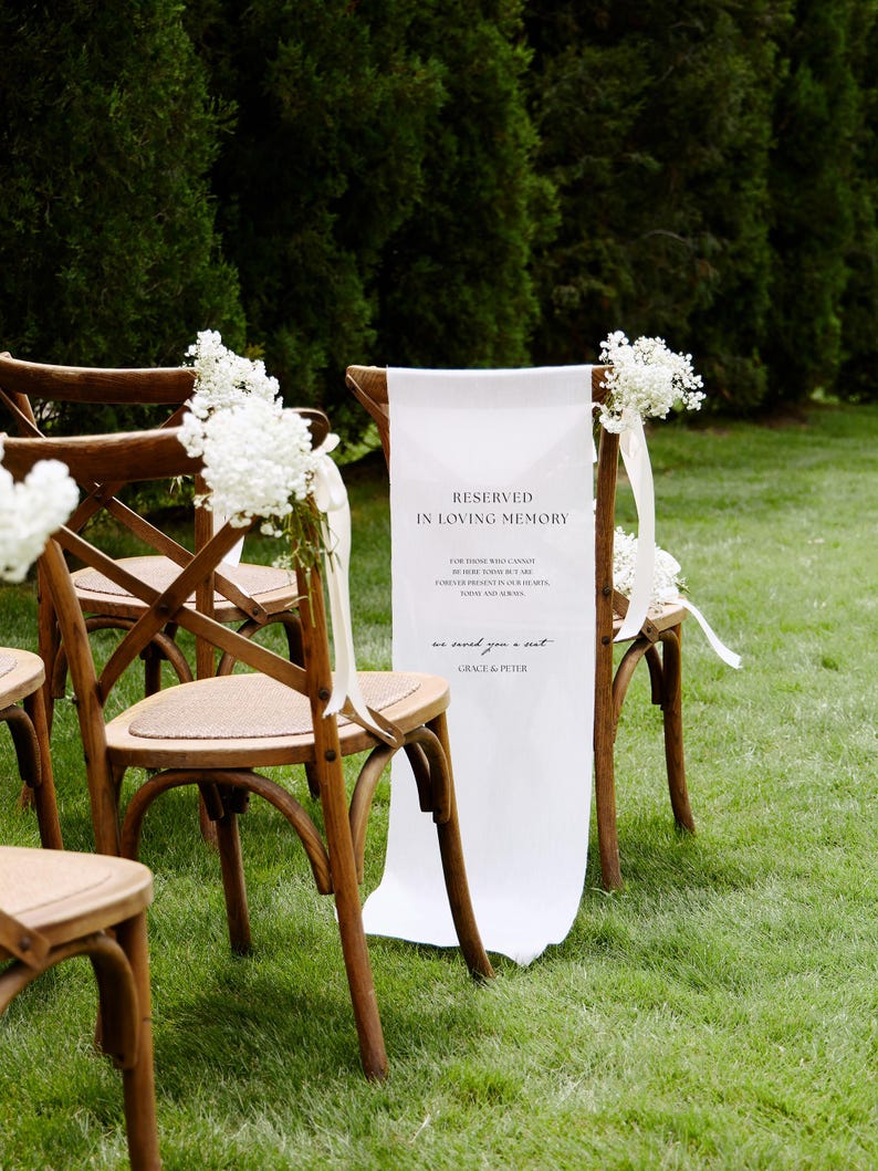 May include: A wooden cross-back chair featuring a white fabric sign draped over the backrest. The sign reads: RESERVED IN LOVING MEMORY, FOR THOSE WHO CANNOT BE HERE TODAY BUT ARE FOREVER PRESENT IN OUR HEARTS, TODAY AND ALWAYS, we saved you a seat, GRACE & PETER. White flowers are tied to the chair.