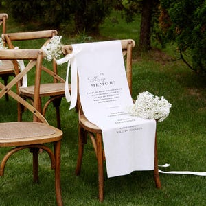May include: A wooden cross-back chair draped with a white fabric memorial sign featuring the text In Loving Memory, followed by a poem and the names Linda Johnson, Sophia Smith, and William Johnson, accented with white baby's breath flowers.
