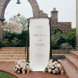 May include: A tall, rectangular white wedding welcome sign featuring black text that reads: WELCOME TO THE WEDDING OF AMELLA and NOAH 13.JULY.2025 TORONTO, CANADA. The sign is mounted on a dark frame with olive green ribbons and floral arrangements.
