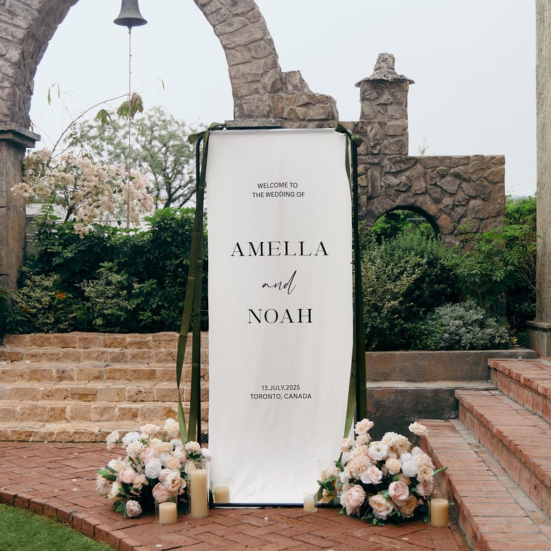 May include: A tall, rectangular white wedding welcome sign featuring black text that reads: WELCOME TO THE WEDDING OF AMELLA and NOAH 13.JULY.2025 TORONTO, CANADA. The sign is mounted on a dark frame with olive green ribbons and floral arrangements.
