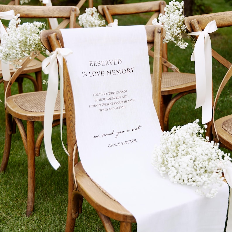 May include: A wooden cross-back chair draped with a white fabric sign featuring black text that reads RESERVED IN LOVE MEMORY, FOR THOSE WHO CANNOT BE HERE TODAY BUT ARE FOREVER PRESENT IN OUR HEARTS, TODAY AND ALWAYS, we saved you a seat, GRACE & PETER.