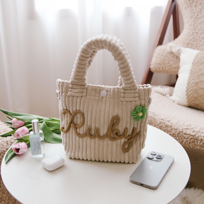 May include: A beige corduroy tote bag with the name "Ruby" embroidered in brown yarn. The bag features a small green flower applique and a button closure. A smartphone, perfume bottle, and wireless earbuds rest on a white table next to the bag.