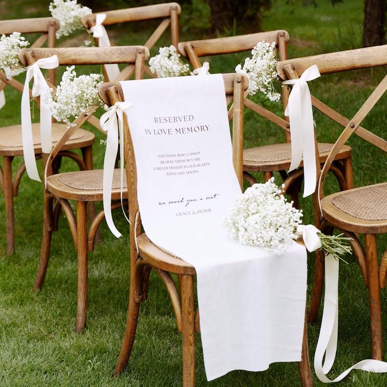 May include: A wooden cross-back chair draped with a white fabric sign reading RESERVED IN LOVE MEMORY, FOR THOSE WHO CANNOT BE HERE TODAY BUT ARE FOREVER PRESENT IN OUR HEARTS, TODAY AND ALWAYS, we saved you a seat, GRACE & PETER, adorned with baby's breath.