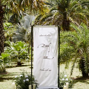 May include: A white wedding sign with black lettering, framed in black, stands in a lush garden. The sign reads "welcome to the wedding of" above "David + Zoe" and "October 25th 2023". The scene is decorated with candles and greenery.