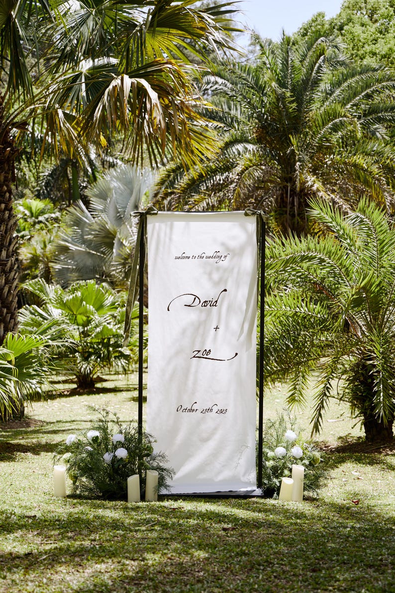 May include: A white wedding sign with black lettering, framed in black, stands in a lush garden. The sign reads "welcome to the wedding of" above "David + Zoe" and "October 25th 2023". The scene is decorated with candles and greenery.